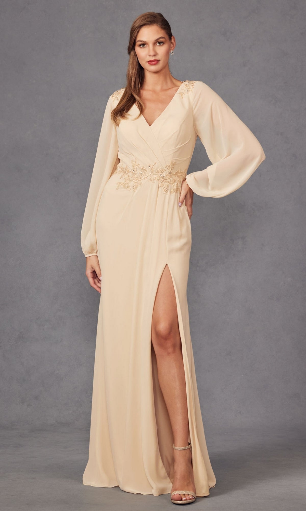 A woman models the Juliet JTM16A V-Neck Long Sleeve Prom Gown with sheer sleeves, floral embroidery at the waist, and a thigh-high slit against a gray background.